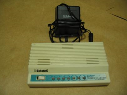 Photograph of a dial-up modem. It is a white, rectangular plastic device. It has a power toggle switch on the top along with seven indicator lights. There is a power cord and plug in the rear of the device.