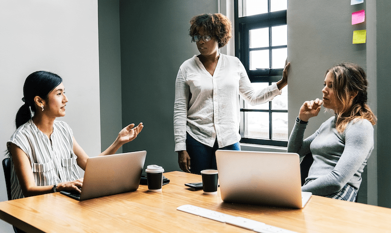 Two women sitting at a table in front of laptops while another woman stands at a whiteboard. All three are having a conversation.